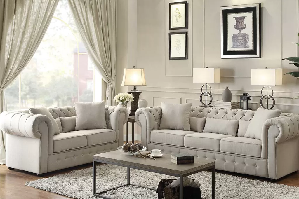 Елегантна вітальня у класичному стилі з м’яким комплектом із дивана та крісла у світлій оббивці, прикрашених каретною стяжкою, декоративними подушками та акцентами в пастельних тонах.