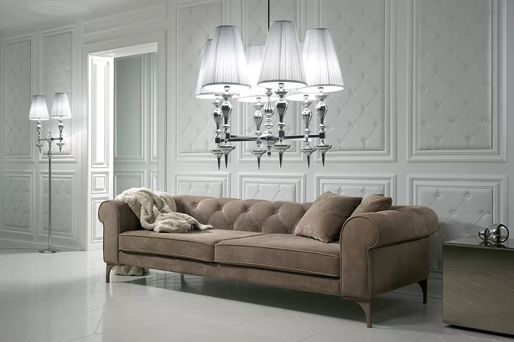 Вишуканий диван у класичному стилі з округлими підлокітниками, м’якими подушками та каретною стяжкою в інтер’єрі з панелями та сріблястими люстрами.