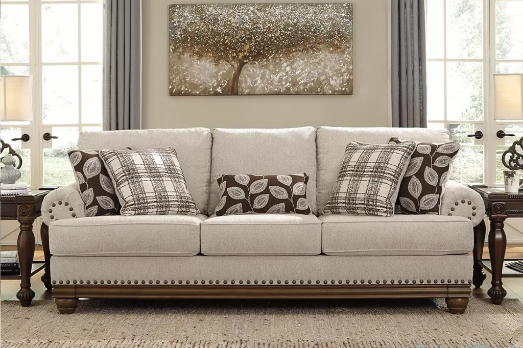 Класичний диван з м’якою текстильною оббивкою, дерев’яним каркасом і подушками з візерунками на фоні великих вікон.