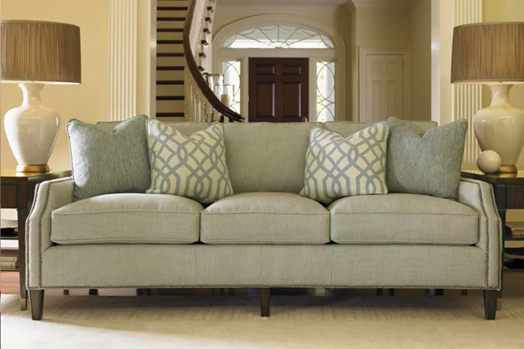 Класичний прямий диван з пастельною оббивкою, дерев’яними ніжками та великими м’якими подушками у просторій вітальні з колонами.